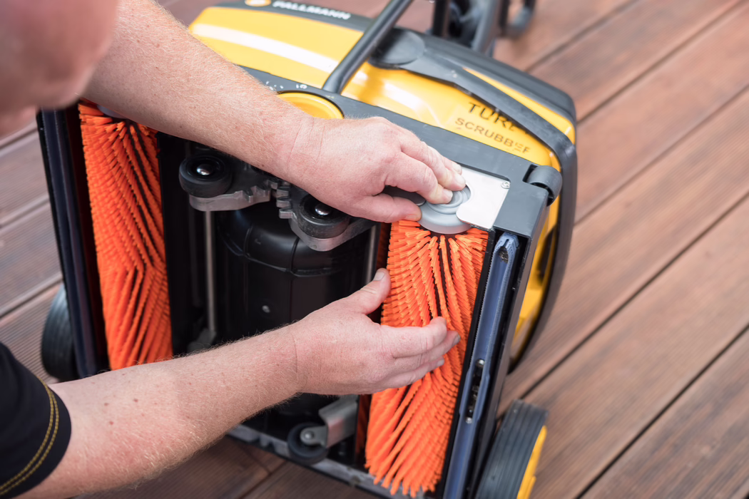 Gros plan sur les mains d'une personne ajustant des brosses cylindriques orange vif sous une machine jaune de lavage de sols posée sur une terrasse en bois.