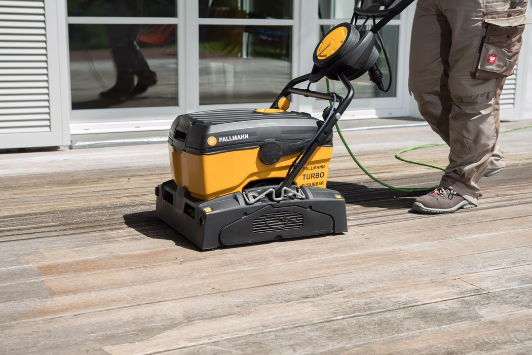 Une monobrosse Pallmann Turbo jaune et noire nettoie une terrasse en bois patinée devant des portes-fenêtres vitrées. À droite, on aperçoit les jambes d'un ouvrier en pantalon de travail brun et chaussures grises ; un câble vert alimente la machine qui laisse une bande nettoyée sur les lames de bois.