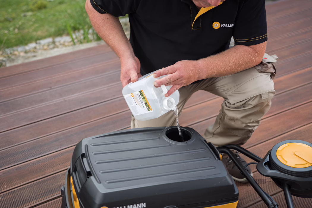 Une personne est agenouillée sur une terrasse en bois et verse du liquide d'un bidon plastique blanc dans l'orifice de remplissage d'un appareil de nettoyage noir et jaune ; elle porte un polo noir aux bordures jaunes avec le logo 'PALLMANN' visible.