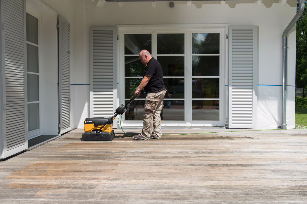 Un homme chauve d'âge moyen, vêtu d'un polo noir et d'un pantalon de travail beige, manœuvre une machine jaune Pallmann pour nettoyer le sol sur une terrasse en bois usée devant une maison blanche. La maison possède de grandes portes vitrées à encadrement blanc avec des petites divisions et des volets à lamelles fermés de chaque côté. La lumière du soleil projette des ombres douces sur la terrasse; le câble d'alimentation vert de la machine traverse les planches en bois; on aperçoit des reflets d'arbres dans les vitrages. La scène illustre un entretien extérieur de routine, avec un soin particulier pour le platelage en bois et un environnement ordonné.