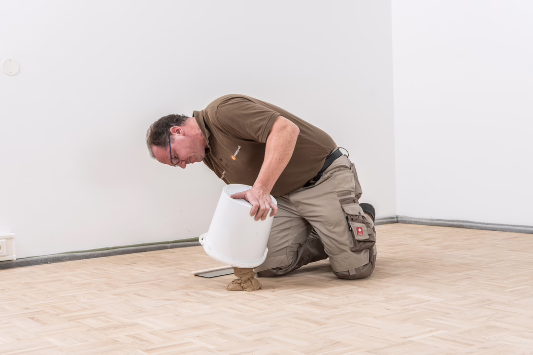 PALLMANN_zudemDSC9098_application image_PALL-X KITT_2017-03 Un ouvrier est agenouillé sur un parquet en bois clair récemment posé dans une pièce aux murs blancs. Il verse avec précaution une masse beiges de colle ou de ragréage depuis un seau en plastique blanc sur le sol en tenant le seau à deux mains; il porte un polo marron, un pantalon de travail beige avec genouillères et poches, et des lunettes.