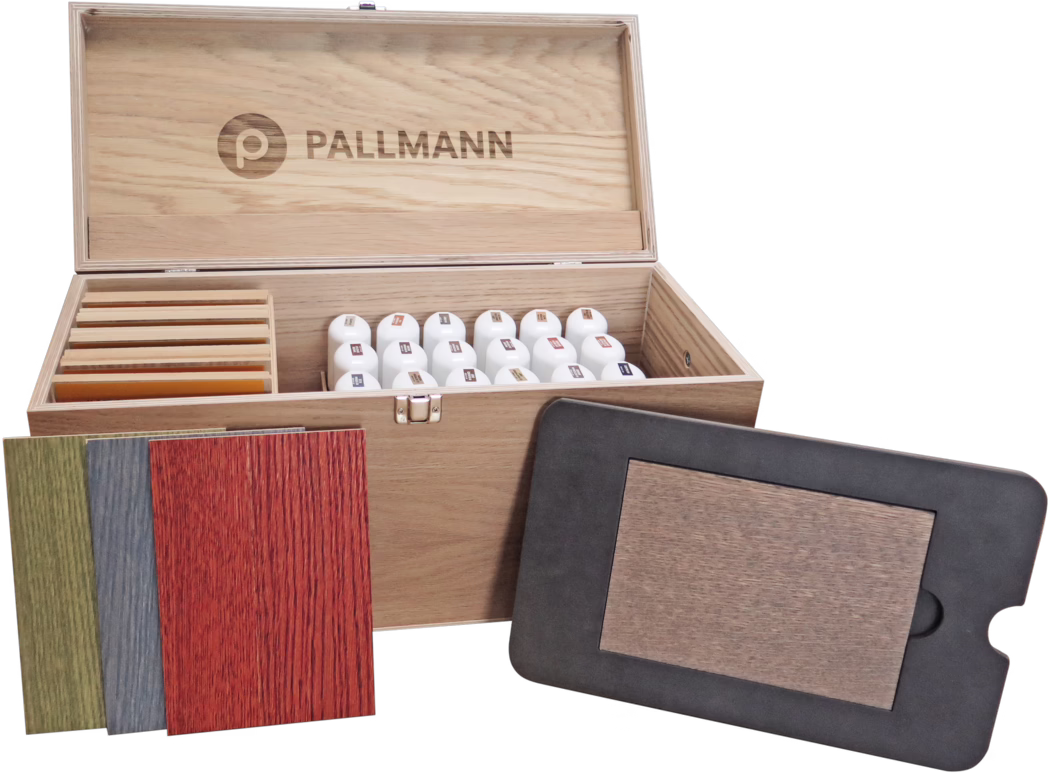 Une mallette de présentation en bois ouverte avec le logo 'PALLMANN' gravé à l'intérieur du couvercle ; l'intérieur contient des rangées de petits flacons blancs et des lames d'échantillons de placage bois organisées. Devant la mallette se trouvent plusieurs échantillons de placage colorés (vert, bleu, rouge) et un échantillon carré encadré de noir. La mallette possède une fermeture métallique et un veinage naturel de chêne.