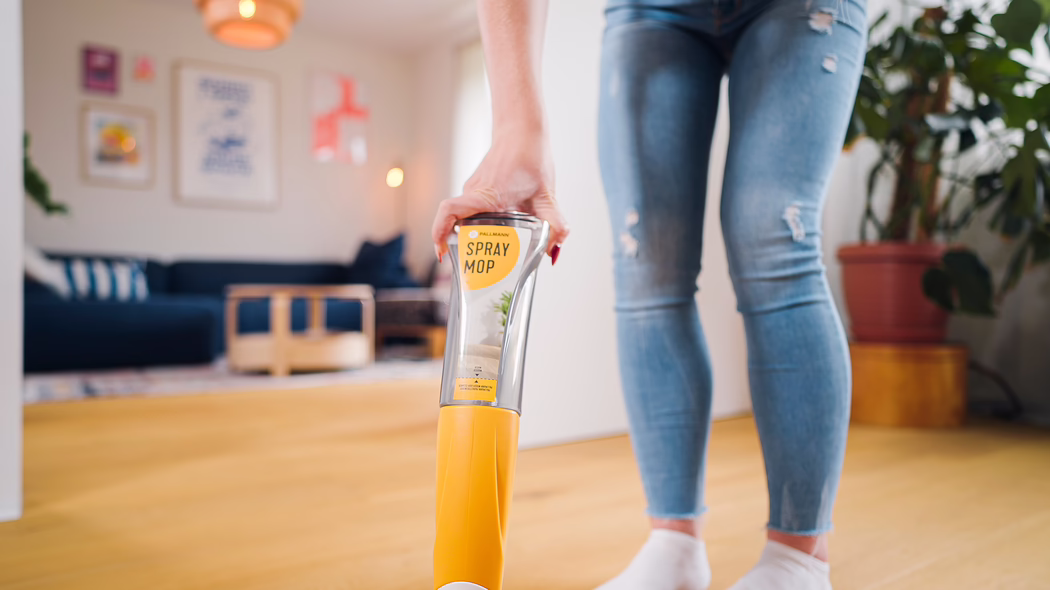 Pallmann_Application Image_Spraymop_072025 (24) Gros plan d'une personne utilisant une vadrouille spray jaune sur un sol en bois clair dans un salon moderne. On voit la poignée et le réservoir transparent du produit nettoyant marqué 'SPRAY MOP', tenu par une main avec du vernis à ongles rouge ; les jambes de la personne portent un jean bleu déchiré et des chaussettes blanches. L'arrière-plan légèrement flou montre un canapé bleu foncé, des cadres muraux, une table basse en bois, une plante en pot et un éclairage chaleureux, créant une ambiance intérieure lumineuse, propre et confortable.